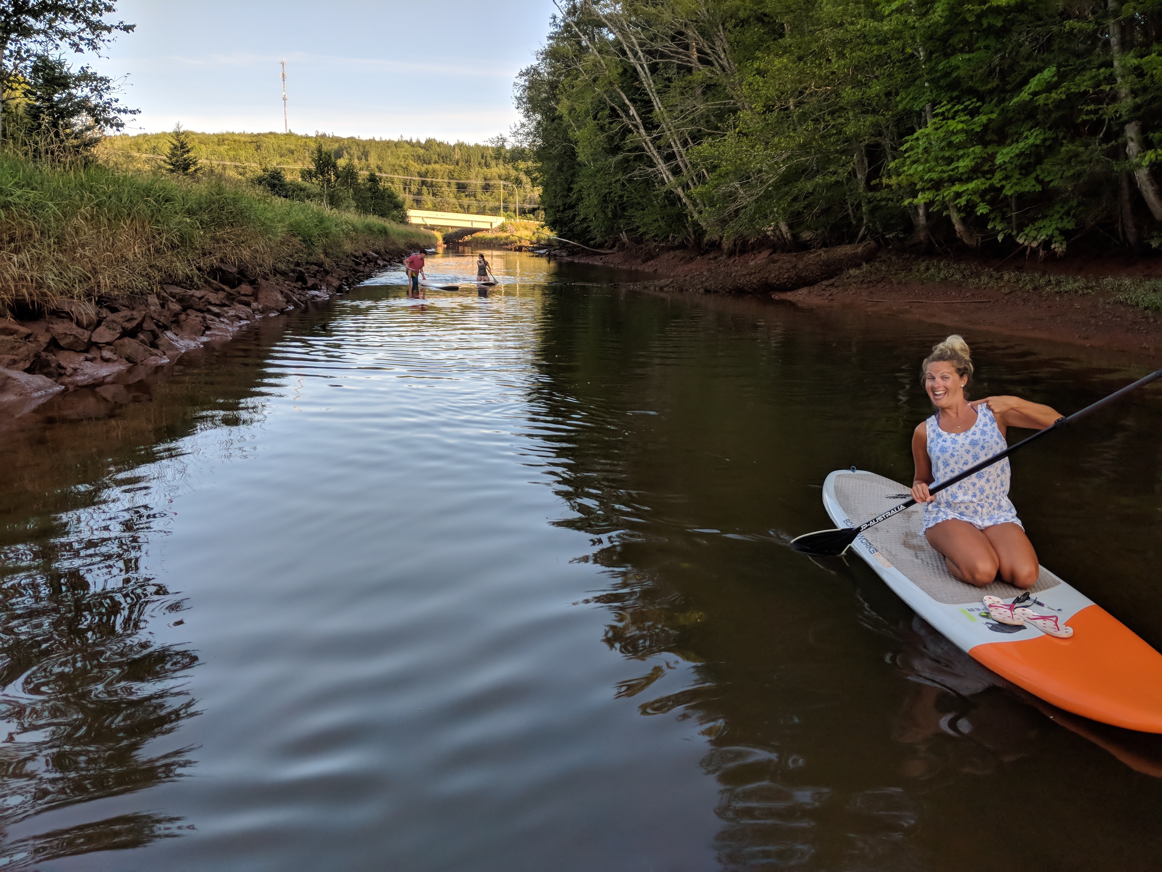 Woman kneeling on a paddleboard on a calm river with trees and a bridge in the background